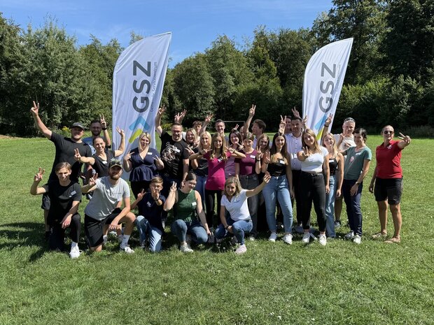 Gruppe von Lehrlingen und Ausbilder:innen des Gemeinde-Servicezentrums bei den Onboarding-Tagen 2025 in Cap Wörth; fröhliche Stimmung im Freien mit GSZ-Fahnen im Hintergrund.